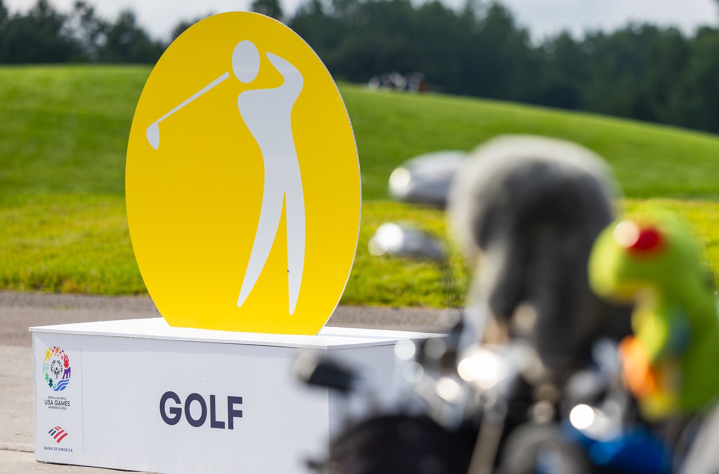 A golf-themed scene with a large yellow sign featuring a white golfer silhouette mounted on a white box labeled GOLF. The background shows a grassy outdoor course with blurred golf carts and a person in the foreground.
