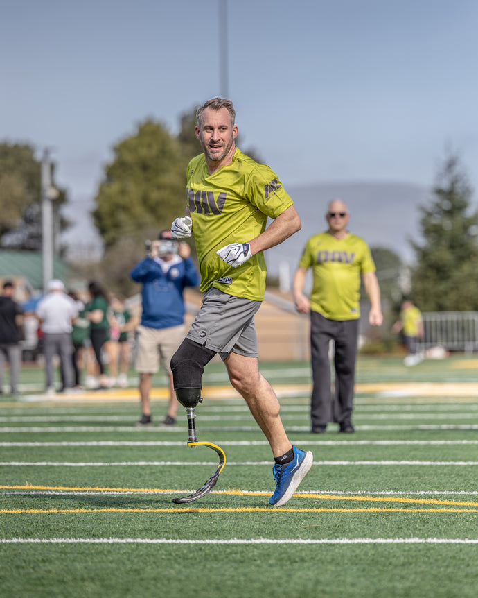 Man with a prosthetic leg running on a sports field with spectators in the background.