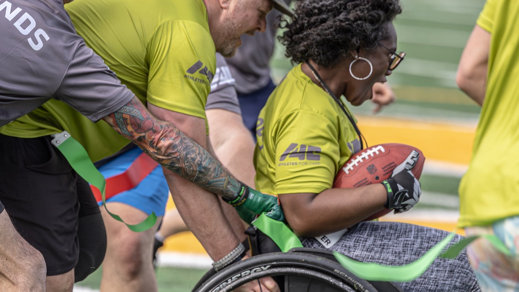 Person in a wheelchair with a football, surrounded by others in sports attire on a field.