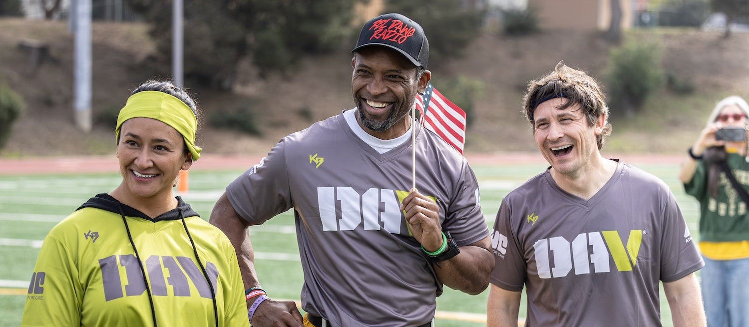 Three people standing together on a sports field, smiling and holding flags.