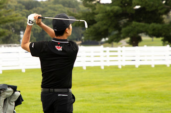 Golfer in black swings club, focusing on the swing with a white fence in the background.