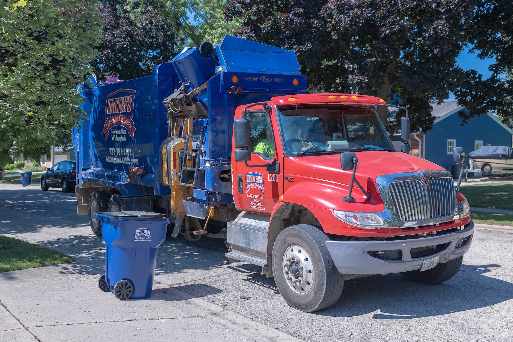 Red and blue garbage truck collecting trash from blue bins on a sunny residential street.