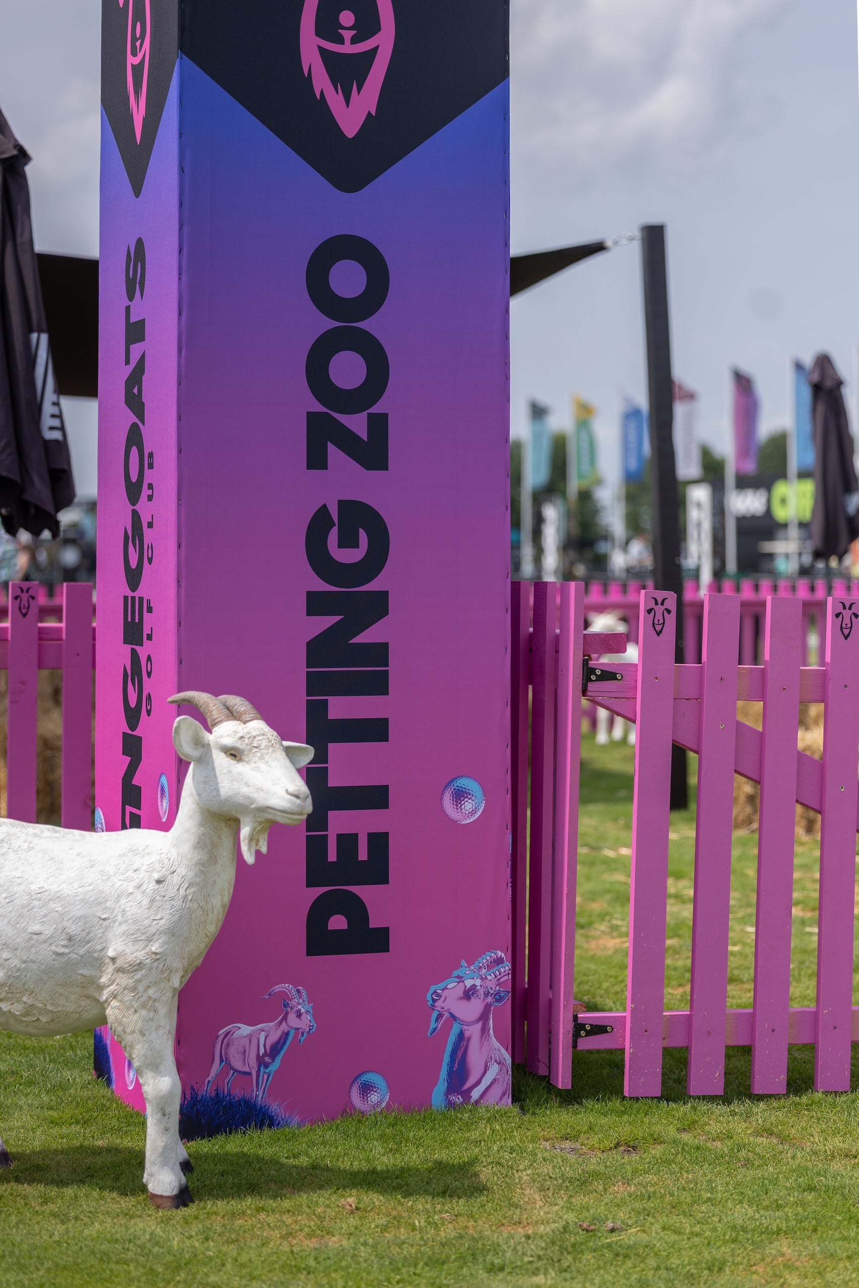 Petting zoo area with a goat statue and pink and purple sign at an outdoor event.