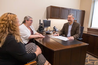 Three people in a professional setting, two sitting at a desk with a third person standing.