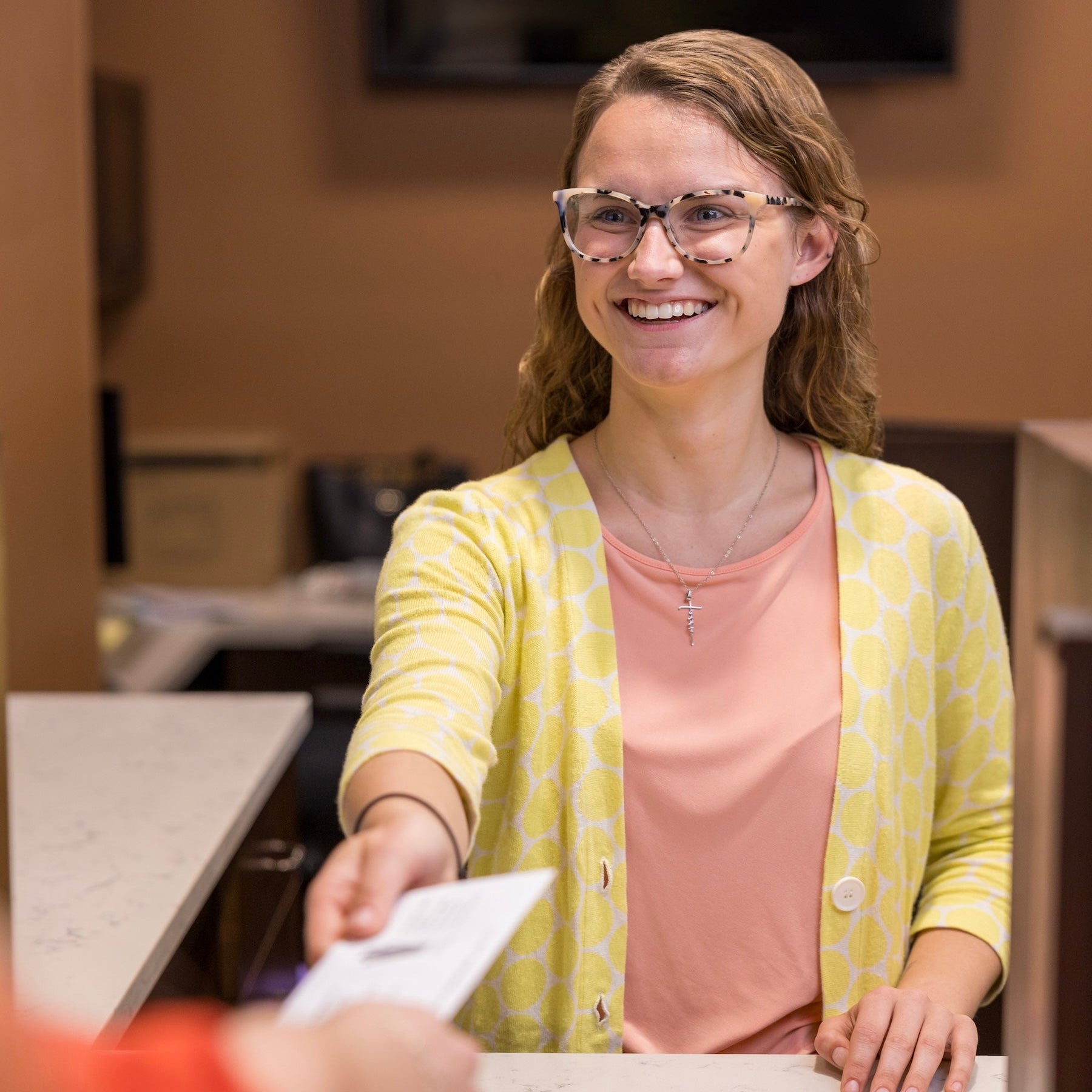 Woman in a yellow cardigan and pink shirt handing over a piece of paper to another person at a counter.