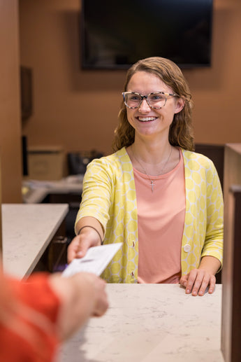 Woman in a yellow cardigan and pink shirt handing over a piece of paper to another person at a counter.