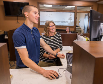 Two people working at a desk with computer monitors in an office setting.