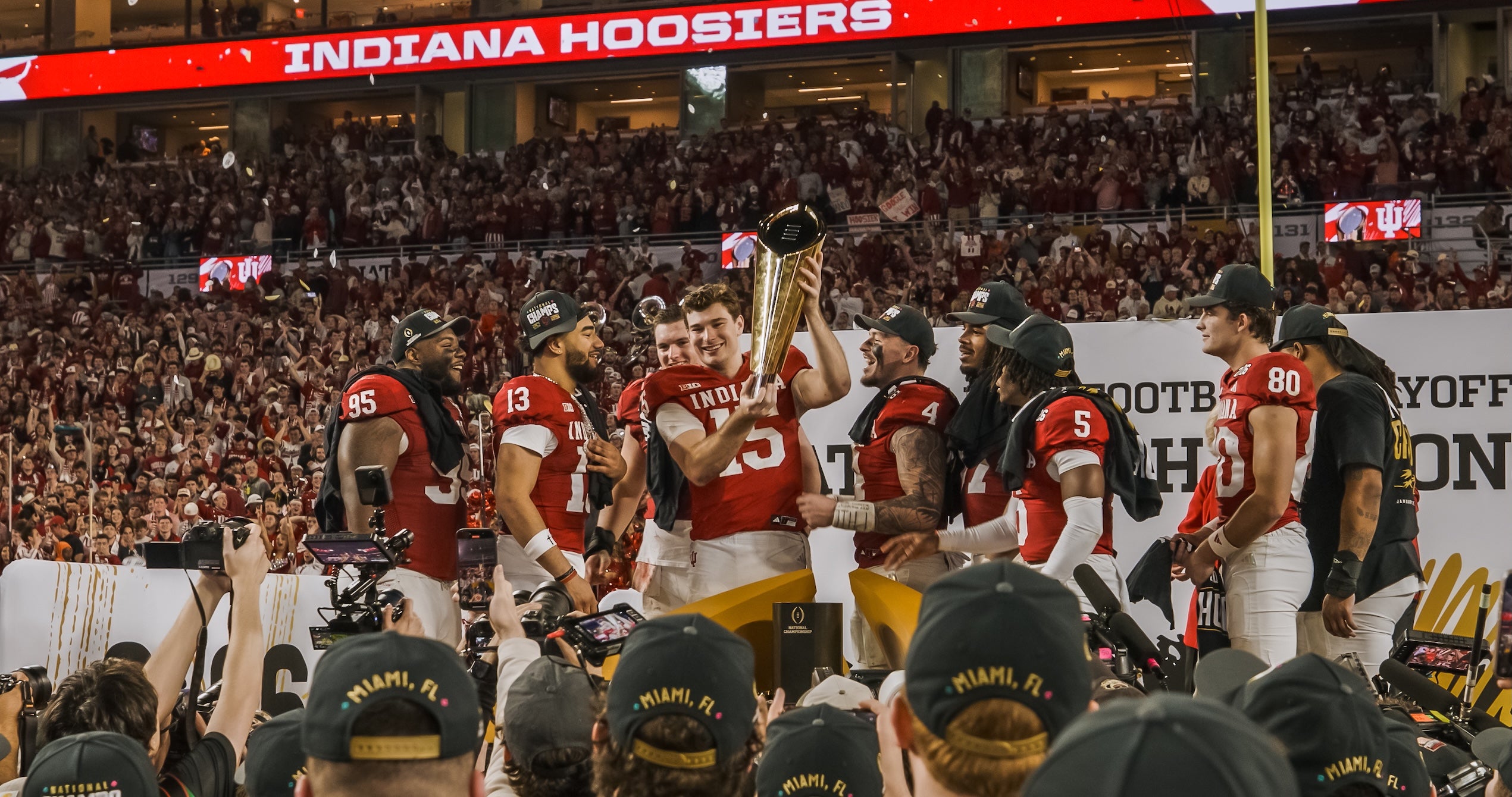 Football players celebrating with a trophy on a field, with 'Indiana Hoosiers' signage in the background.