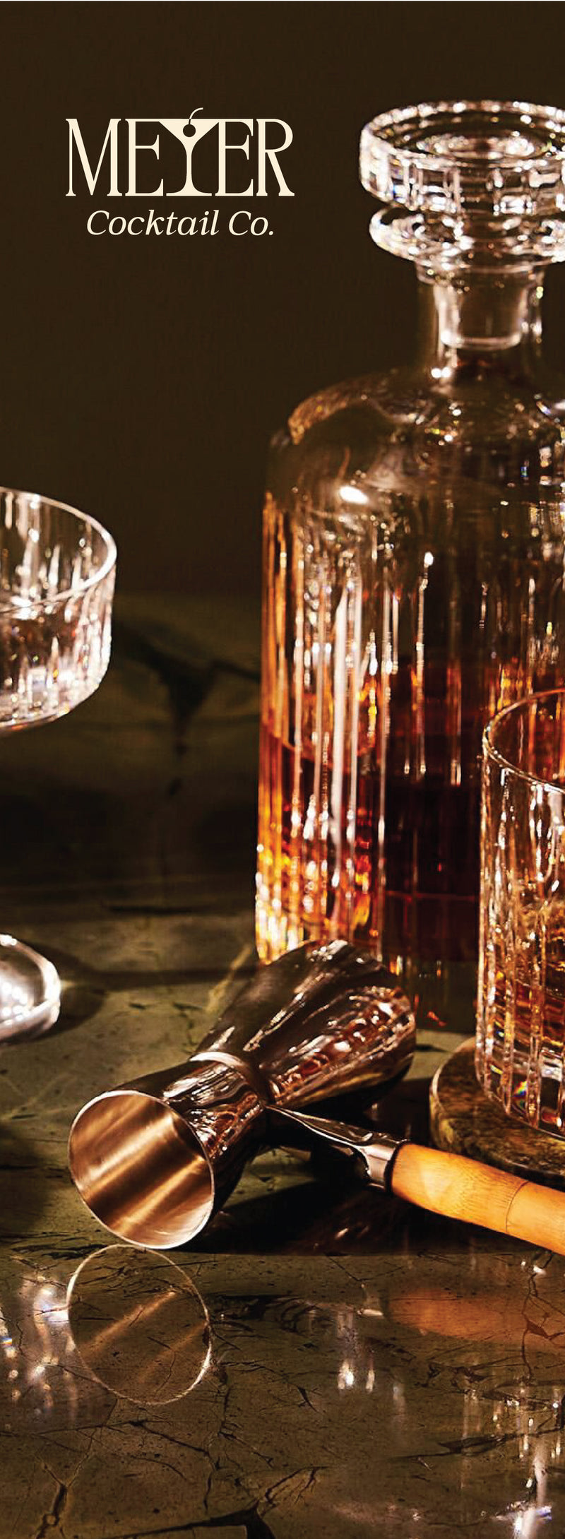 Cocktail setup with MEYER Cocktail Co. branding, including a decanter, glasses, and bar tools on a reflective surface.