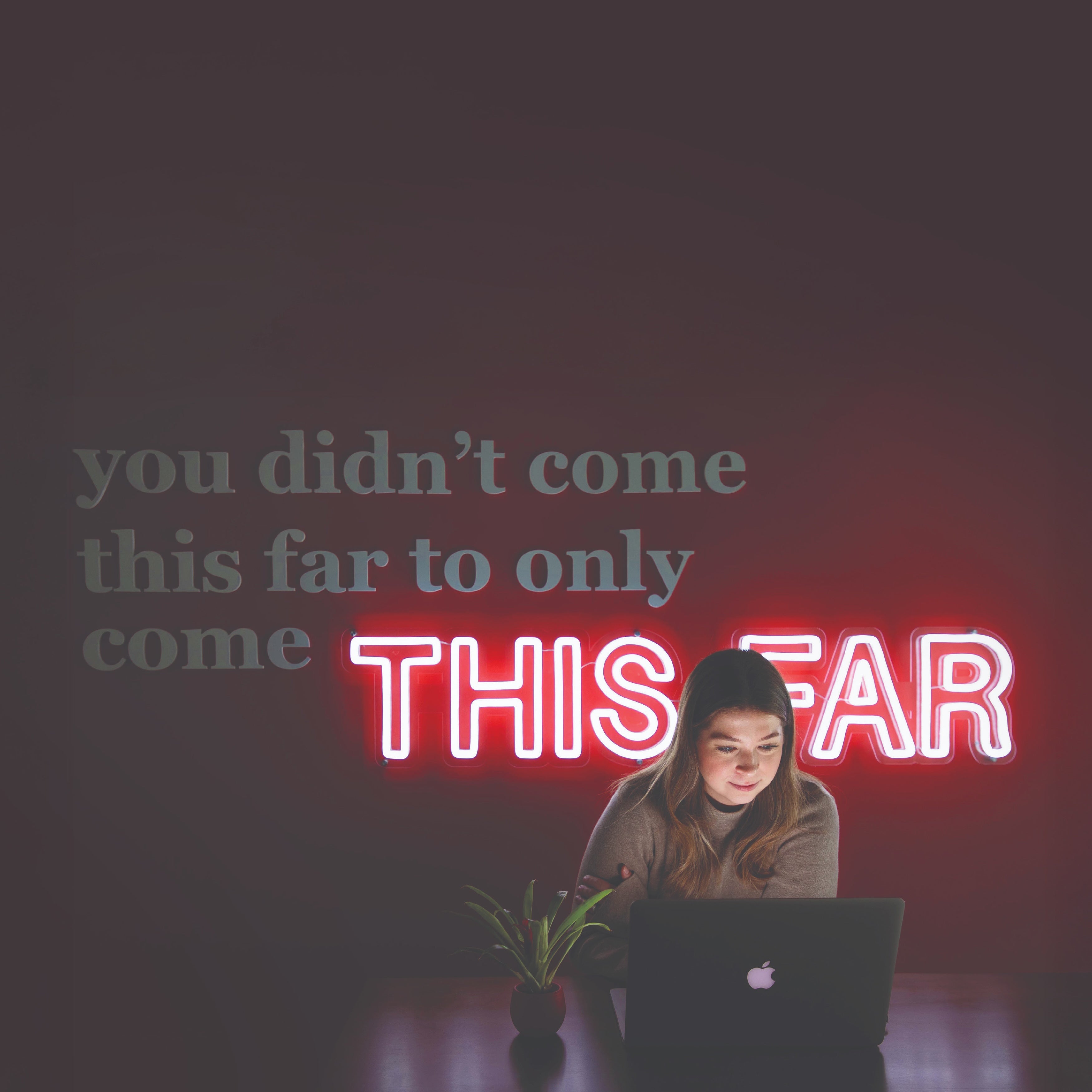 Woman working on laptop in front of neon sign "you didn't come this far to only come THIS FAR".
