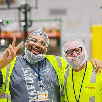Two individuals in a warehouse setting, wearing safety vests and hairnets.