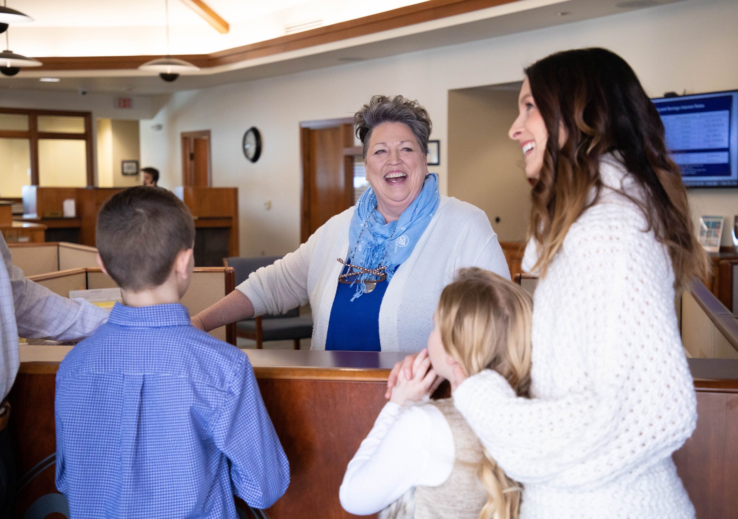 Smiling woman with blue scarf laughs with a family in a bank lobby.