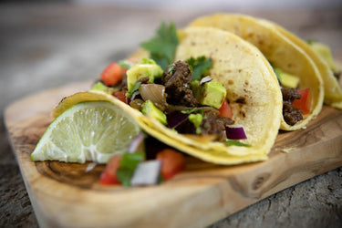 Tacos with beef, vegetables, and avocado on a wooden board with a lime wedge.