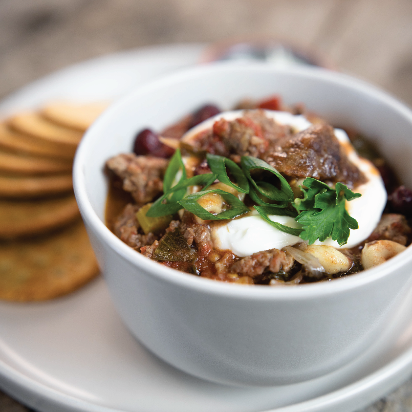 White bowl of chili with sour cream and green onions on a wooden surface