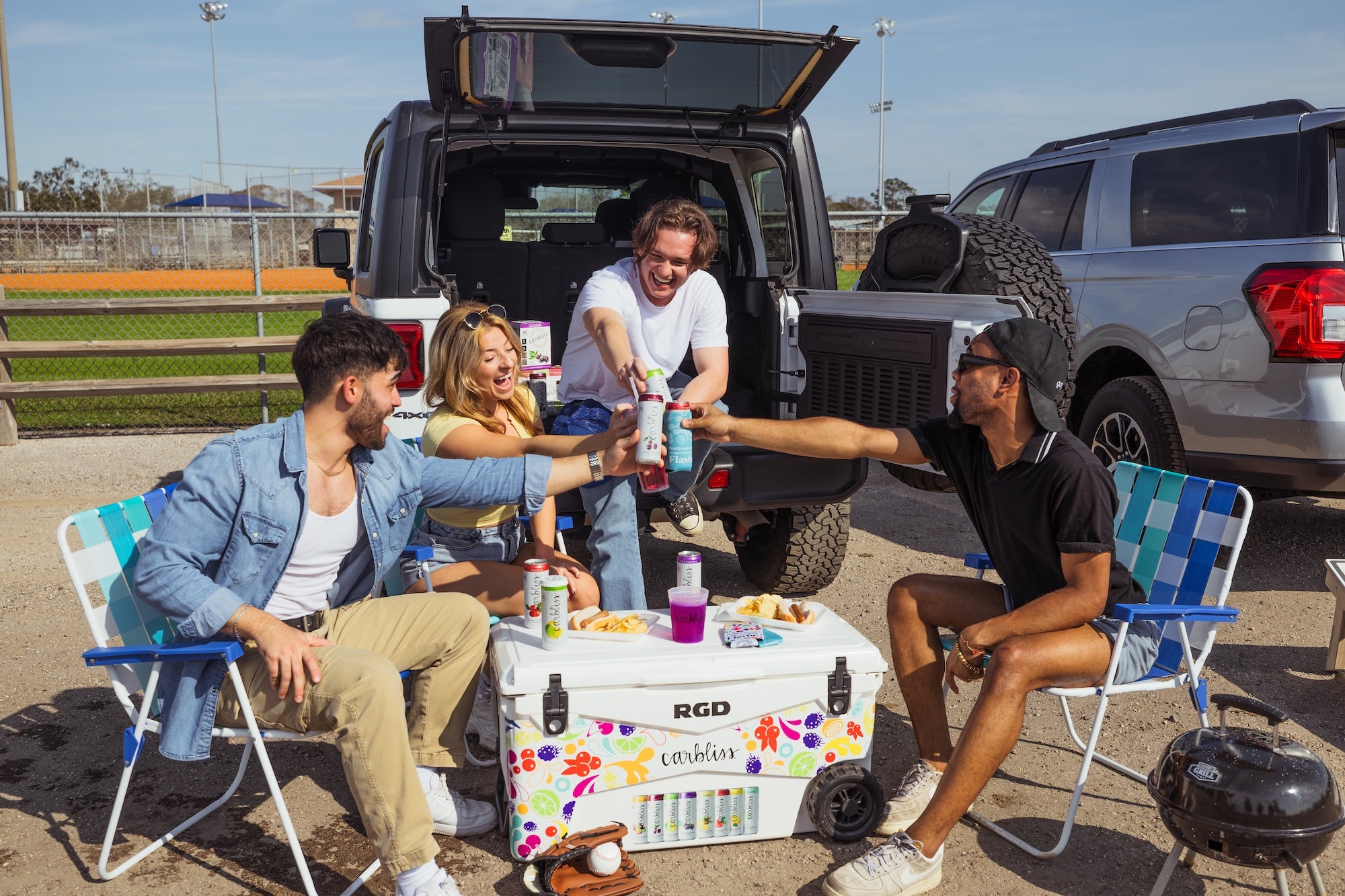 Friends toasting drinks around a cooler at a tailgate party with two SUVs and a baseball field in background.