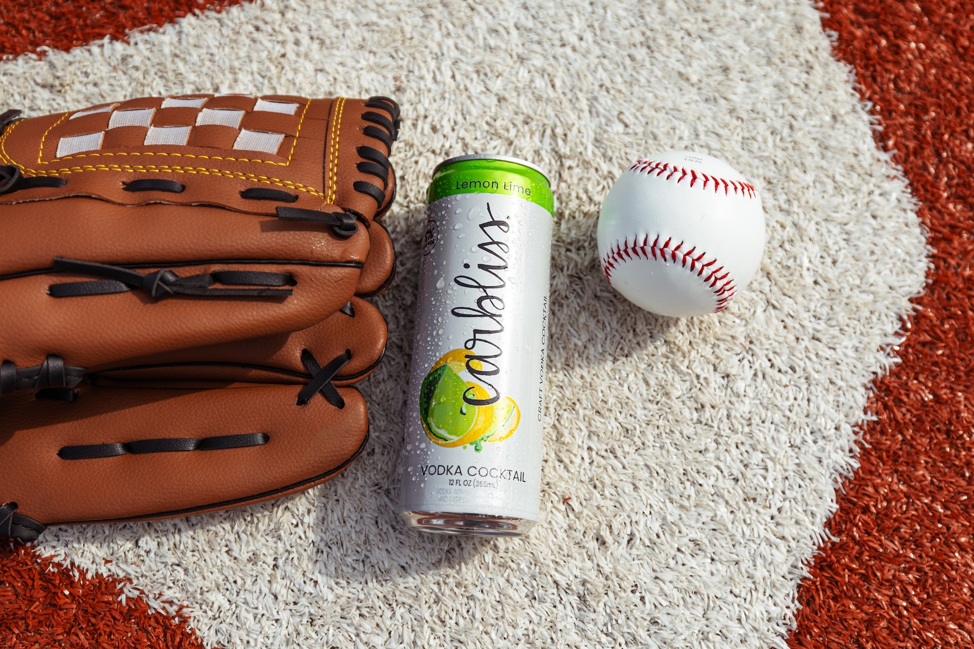 Baseball glove and ball with a can of "carbliss" lemon lime vodka cocktail on a textured surface.