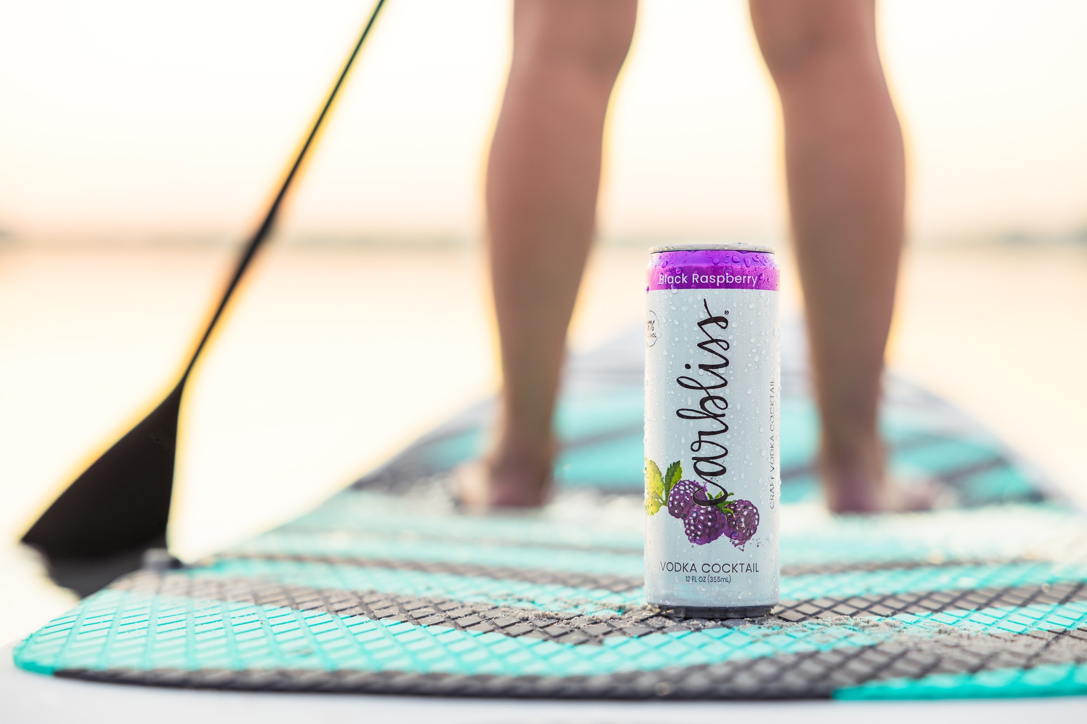 Can of Black Raspberry Vodka Cocktail on a paddleboard with feet in the background at sunset.