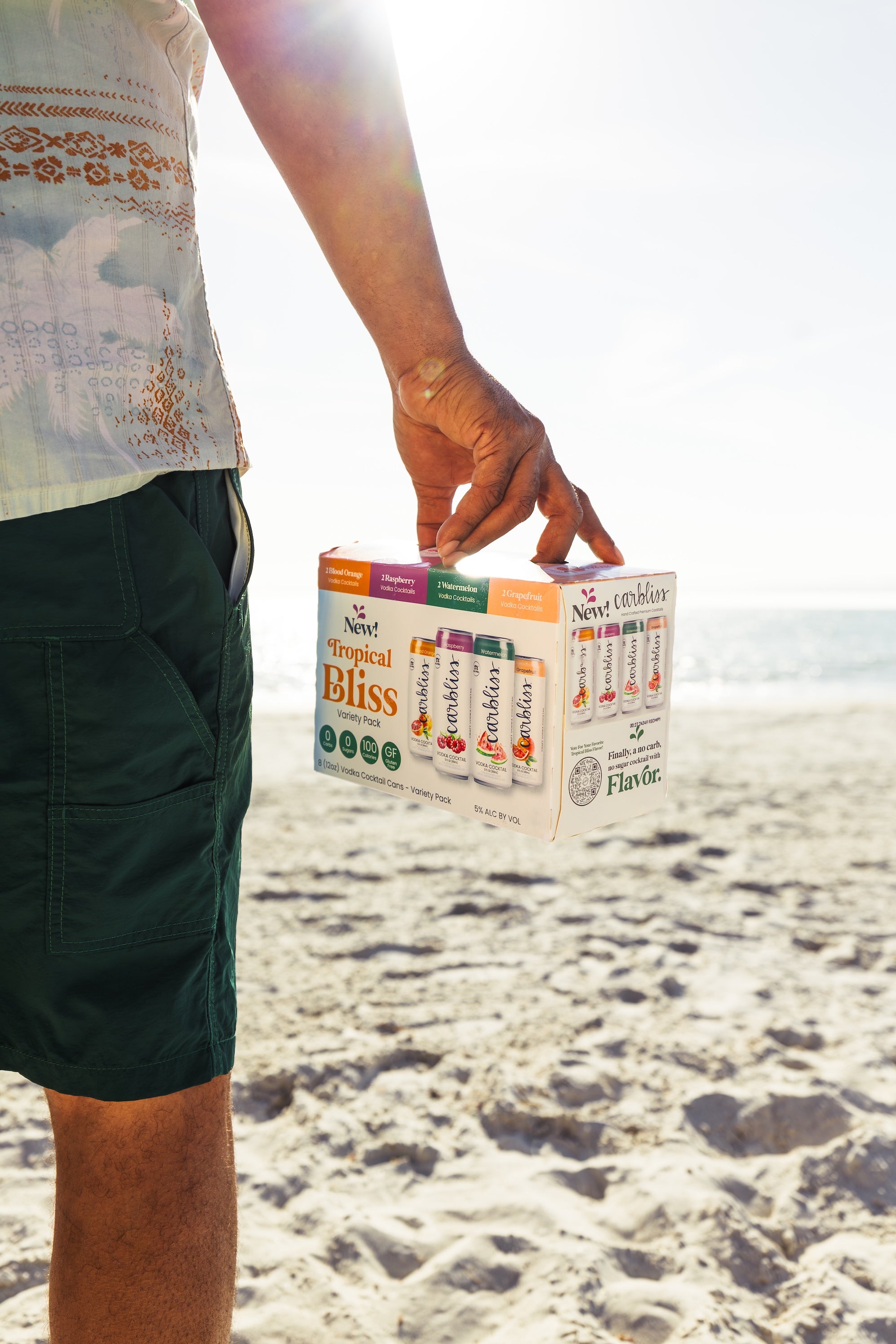 Man holds a variety pack of Carbliss Tropical Bliss vodka cocktails on a sunny beach.