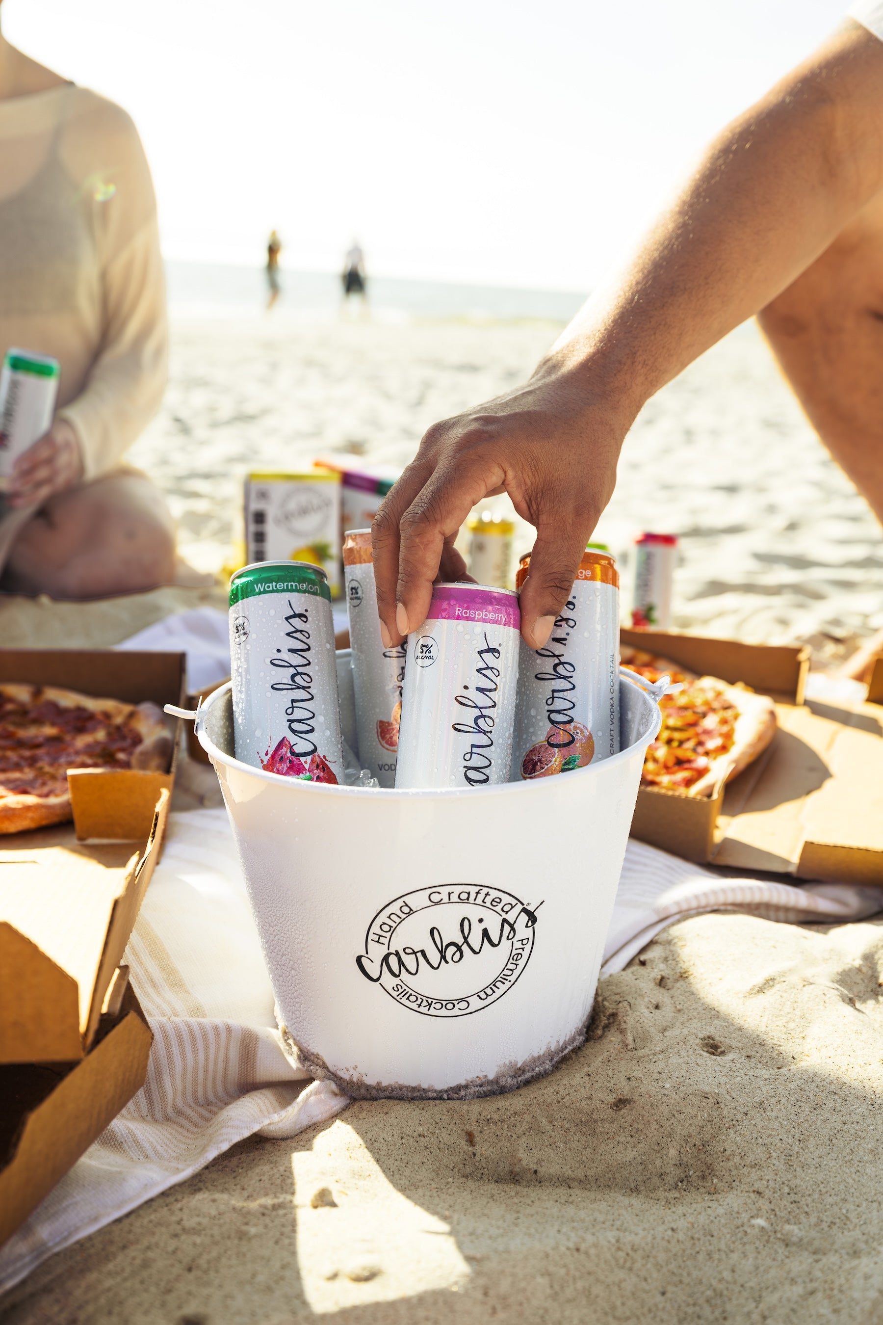 Hand reaching into a chilled bucket of Carbliss cocktails on a beach, with pizza in the background.