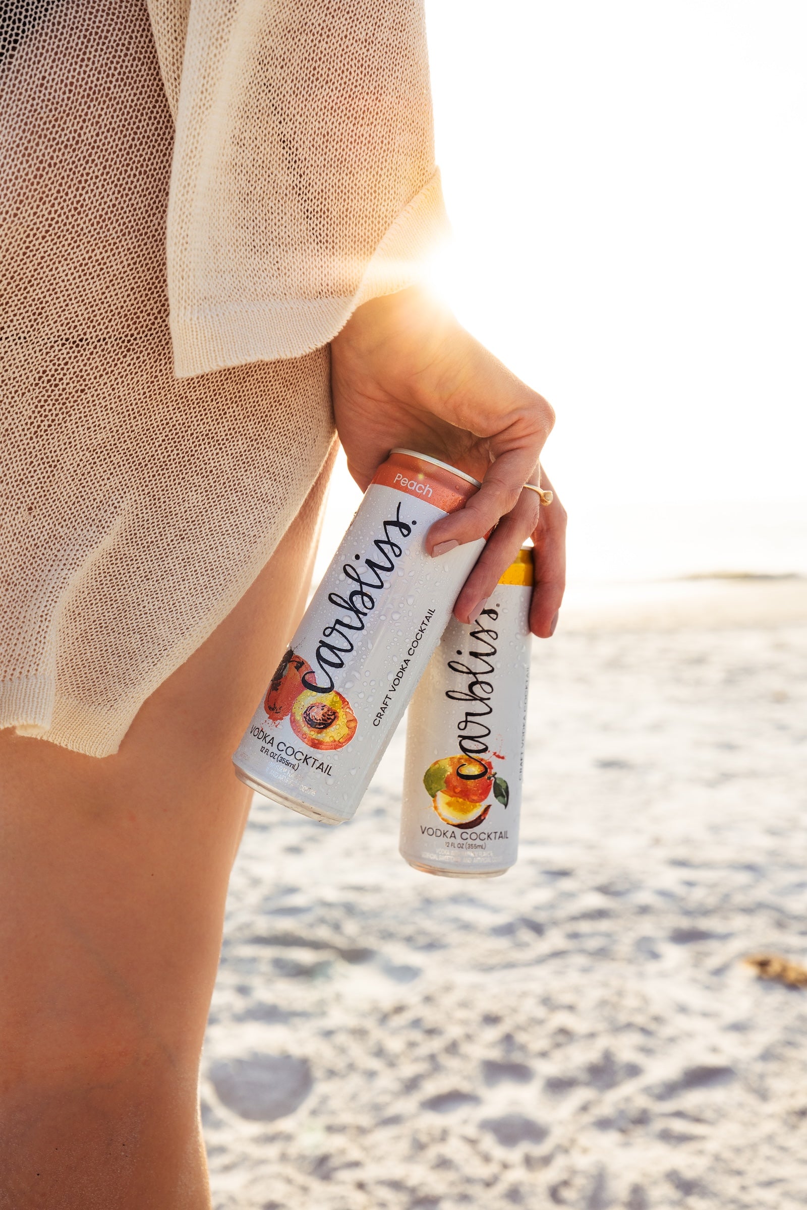 Woman holding two peach vodka cocktail cans on a sandy beach at sunset.