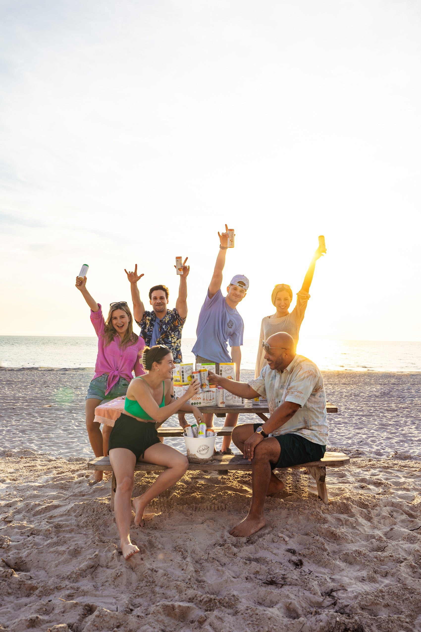Group of friends toasting drinks on a beach at sunset with a cooler and cases of drinks.