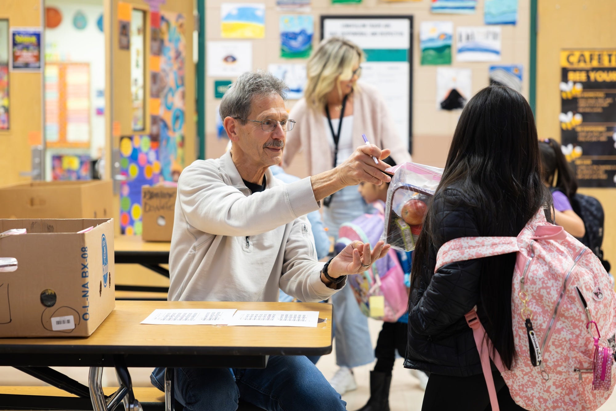 Teacher hands a lunch bag to a student with a pink floral backpack at school.