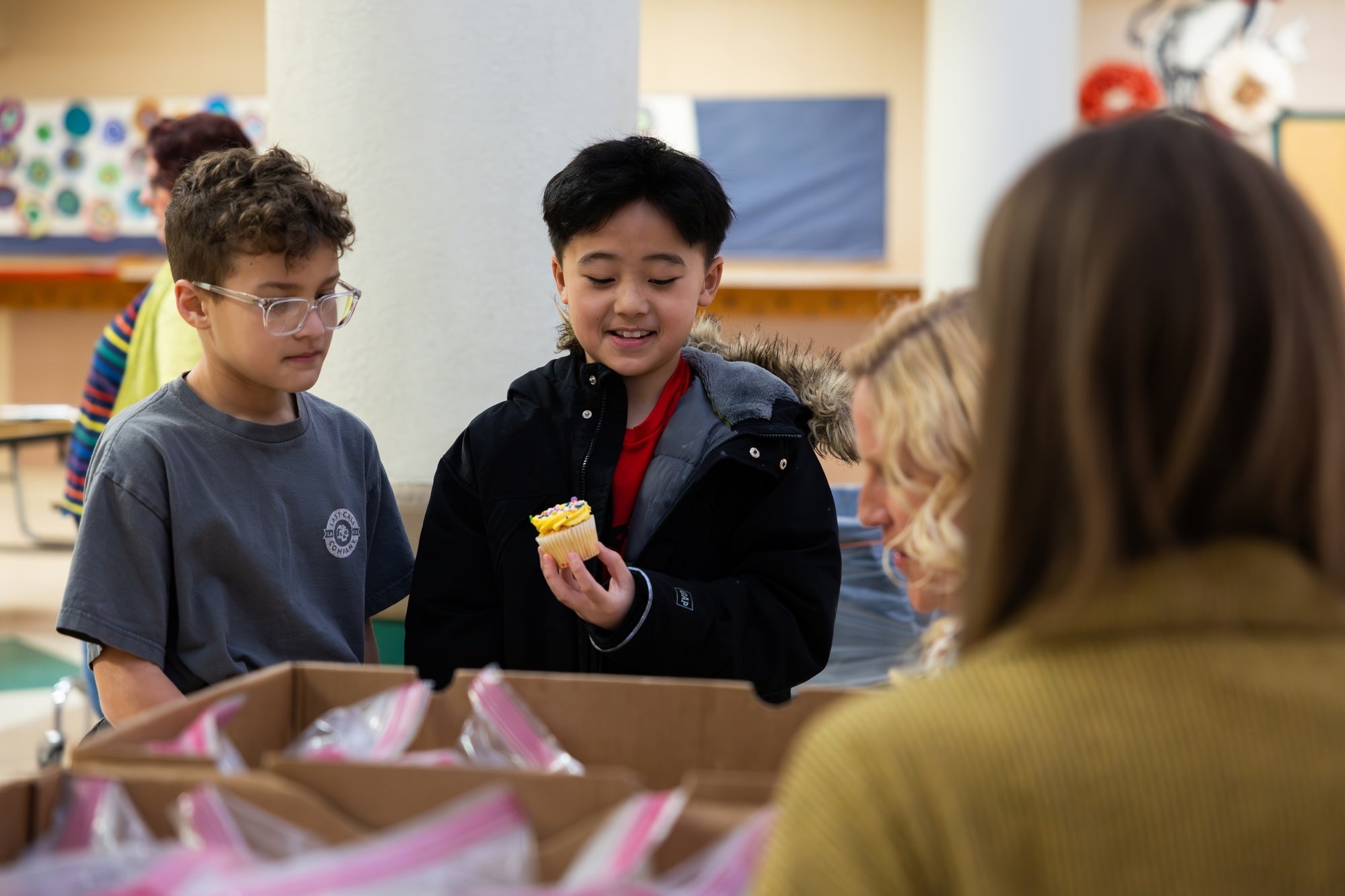 Boy holds a cupcake while a diverse group prepares food boxes.