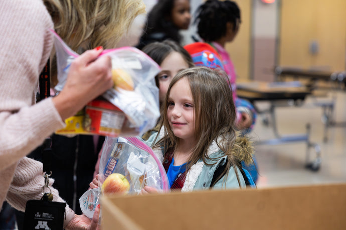 A person hands a child a bagged lunch containing an apple and yogurt.
