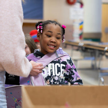 Young Black girl with pink beads in her hair smiles while having a backpack adjusted.