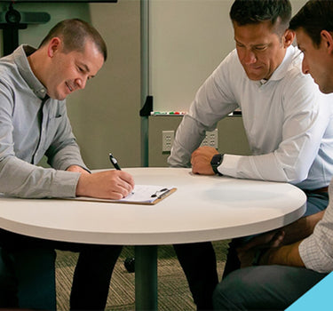 Three men in a meeting at a round table. One man writes on a clipboard while the others watch.