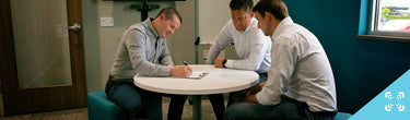 Three men in a modern office meeting around a round table, one writing on a clipboard.