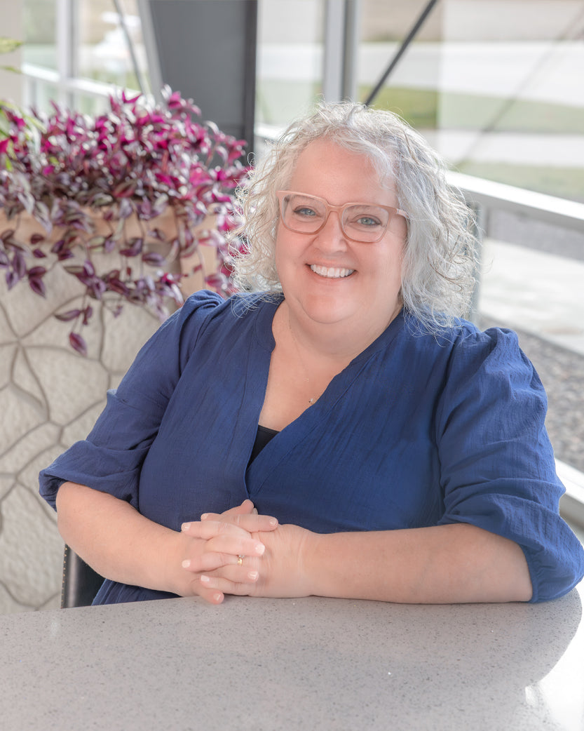 Woman with curly gray hair and glasses smiles, hands clasped, wearing a blue top.