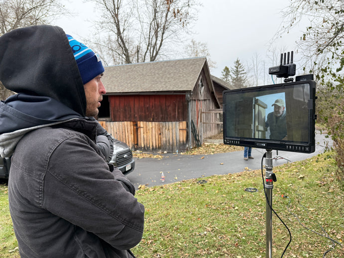 Person operating a camera with a monitor outdoors, near a wooden building.