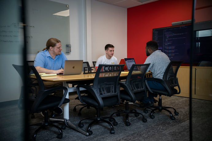 Three men in a modern office meeting room, collaborating around a table with laptops and a whiteboard.