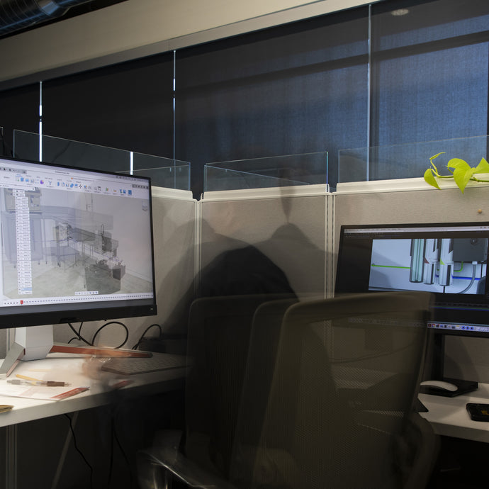 Office desk with dual monitors showing CAD software and pipes, with a blurred figure seated.