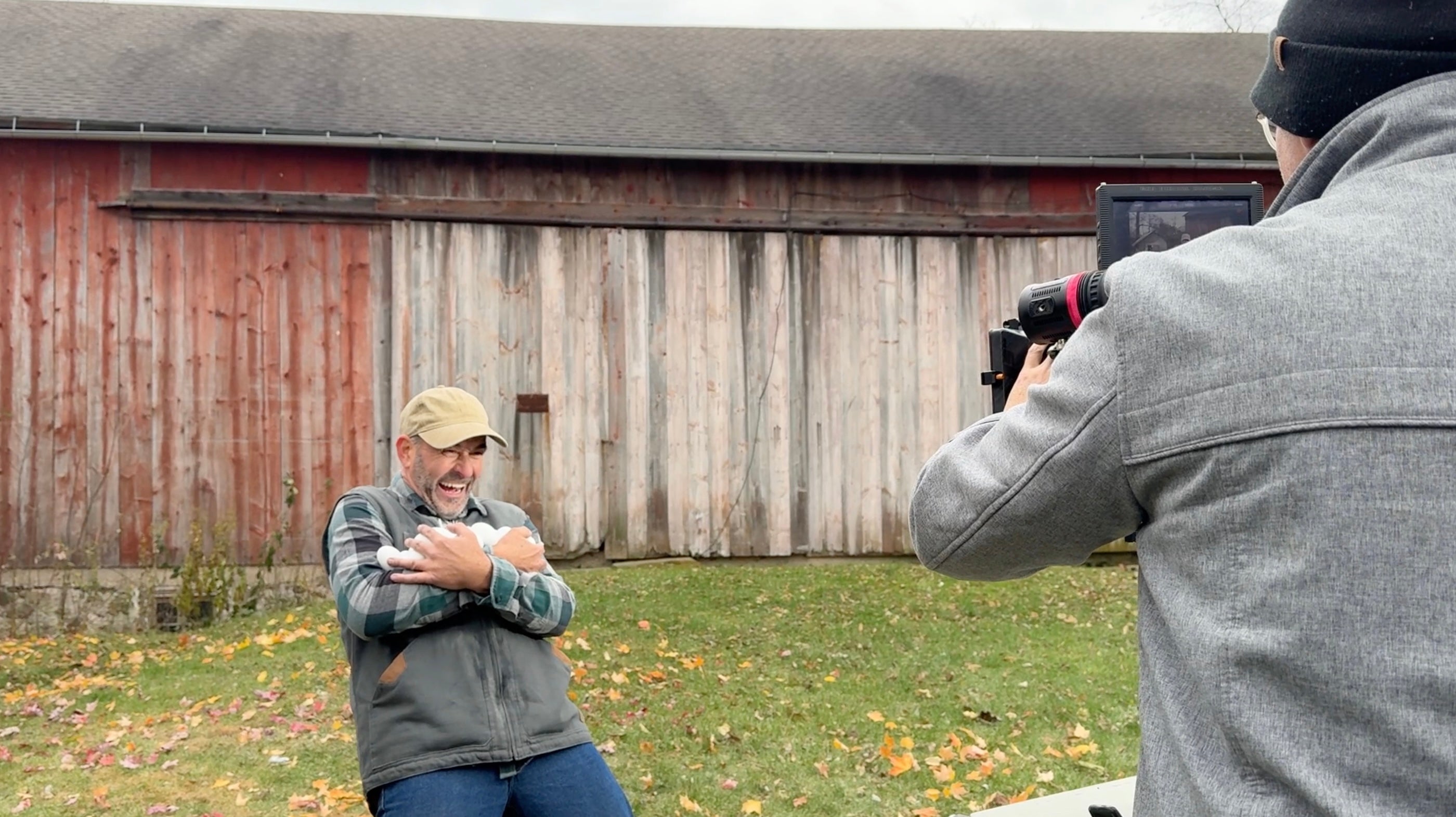Person holding a baby in front of a barn with another person taking a photo.