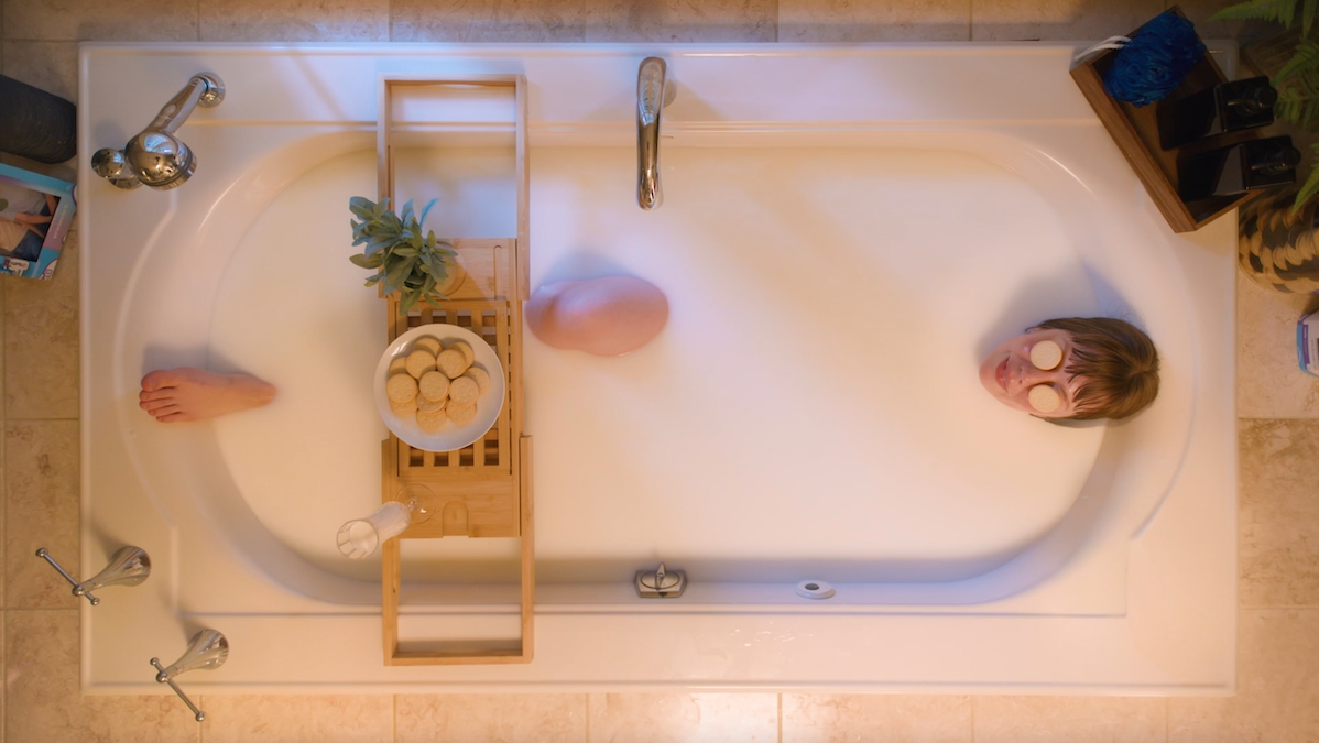Overhead view of a person relaxing in a bubble bath, wearing spa eye masks. A wooden caddy holds a bowl of bath bombs and a small plant, with a pink bath bomb floating in the tub.