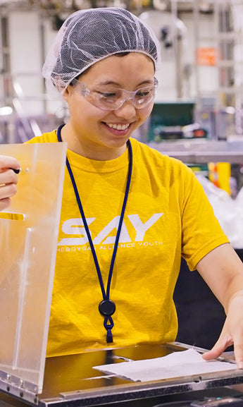 Person wearing a yellow shirt with 'SAY' logo in a factory setting