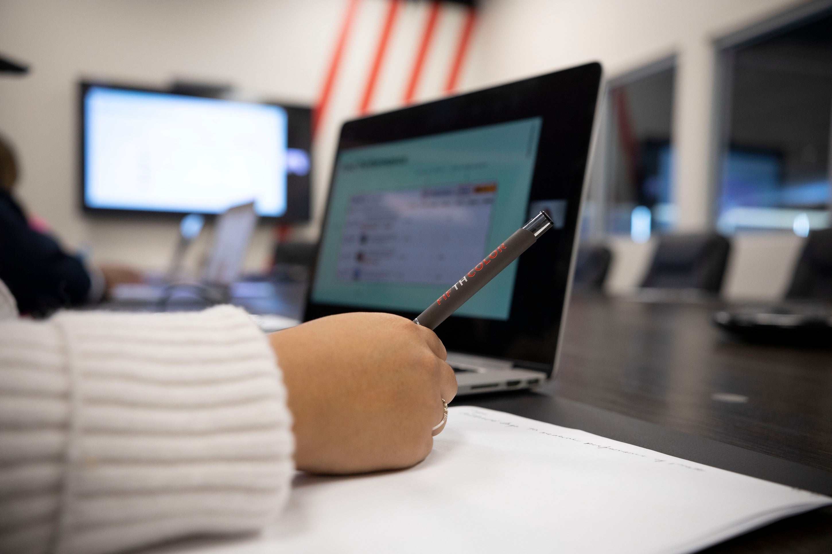 Person's hand with a pen writing on paper, laptop in background.