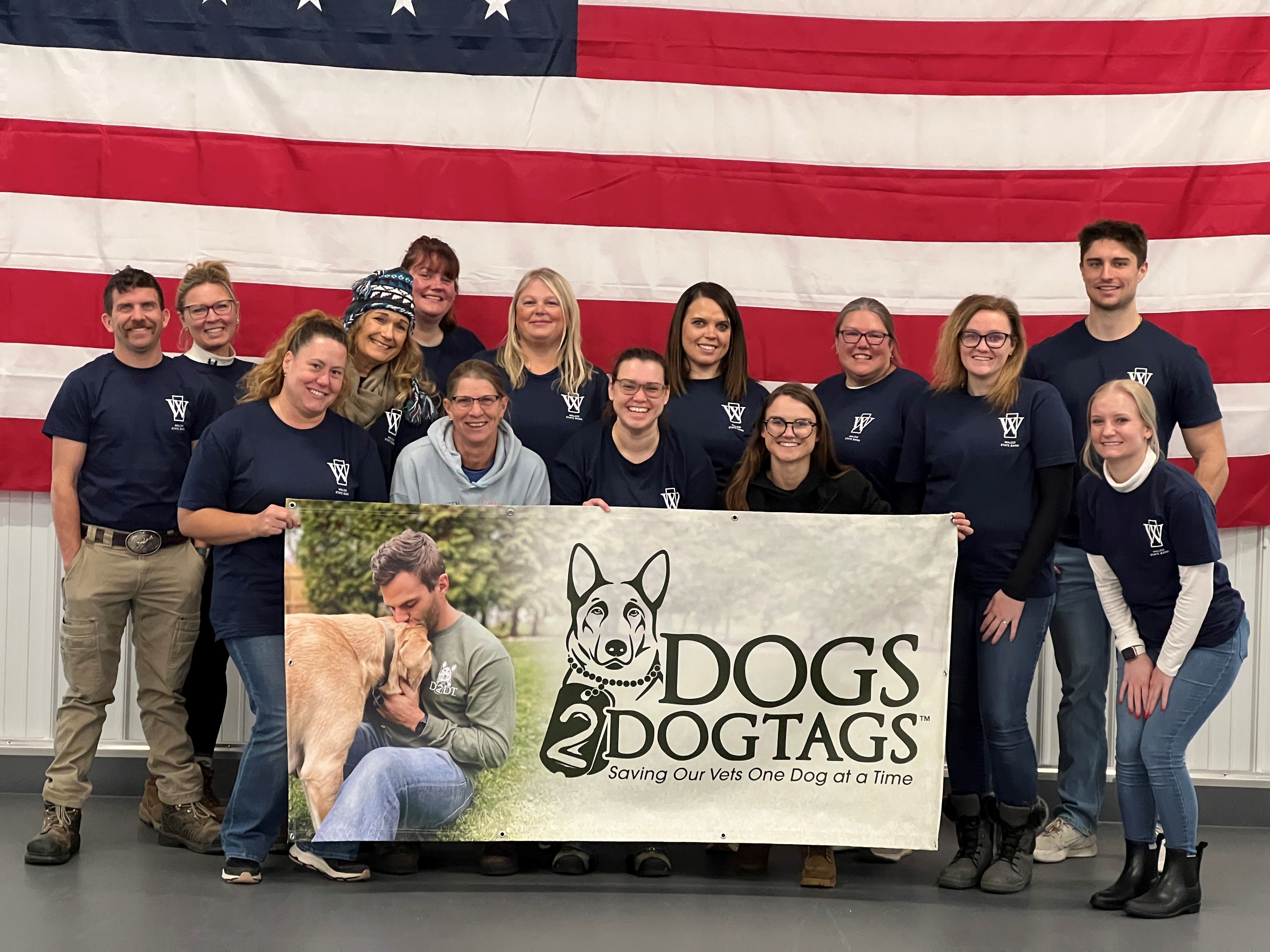 Group of people in matching shirts holding a 'DOGS 2 DOGTAGS' sign in front of an American flag.