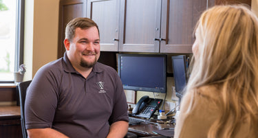 Man in a gray shirt sitting at a desk with a woman, smiling and engaged in conversation.