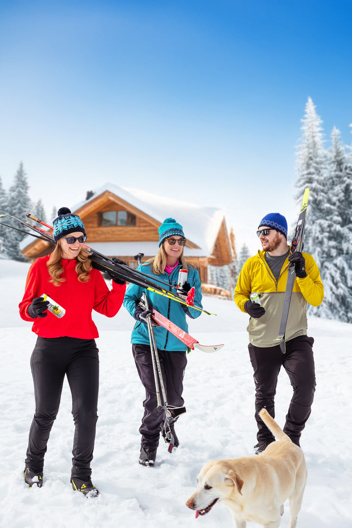 Three people with skis and a dog in a snowy landscape with a wooden cabin.