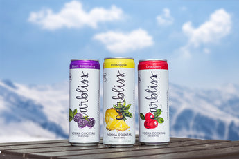 Three cans of Ardbellus vodka cocktails on a wooden surface with a blurred outdoor background.