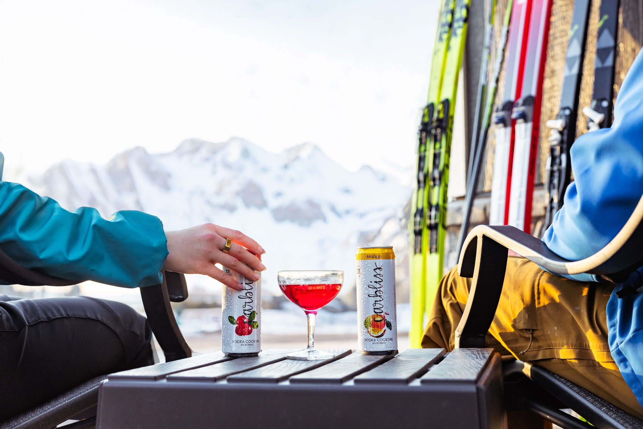 Two people sitting at a table with drinks and skis in the background, mountains visible