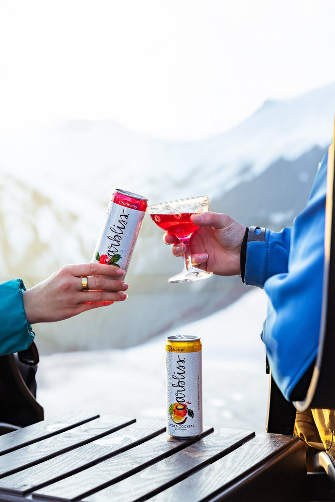 Two people toasting with a can and a glass of red liquid against a mountainous background