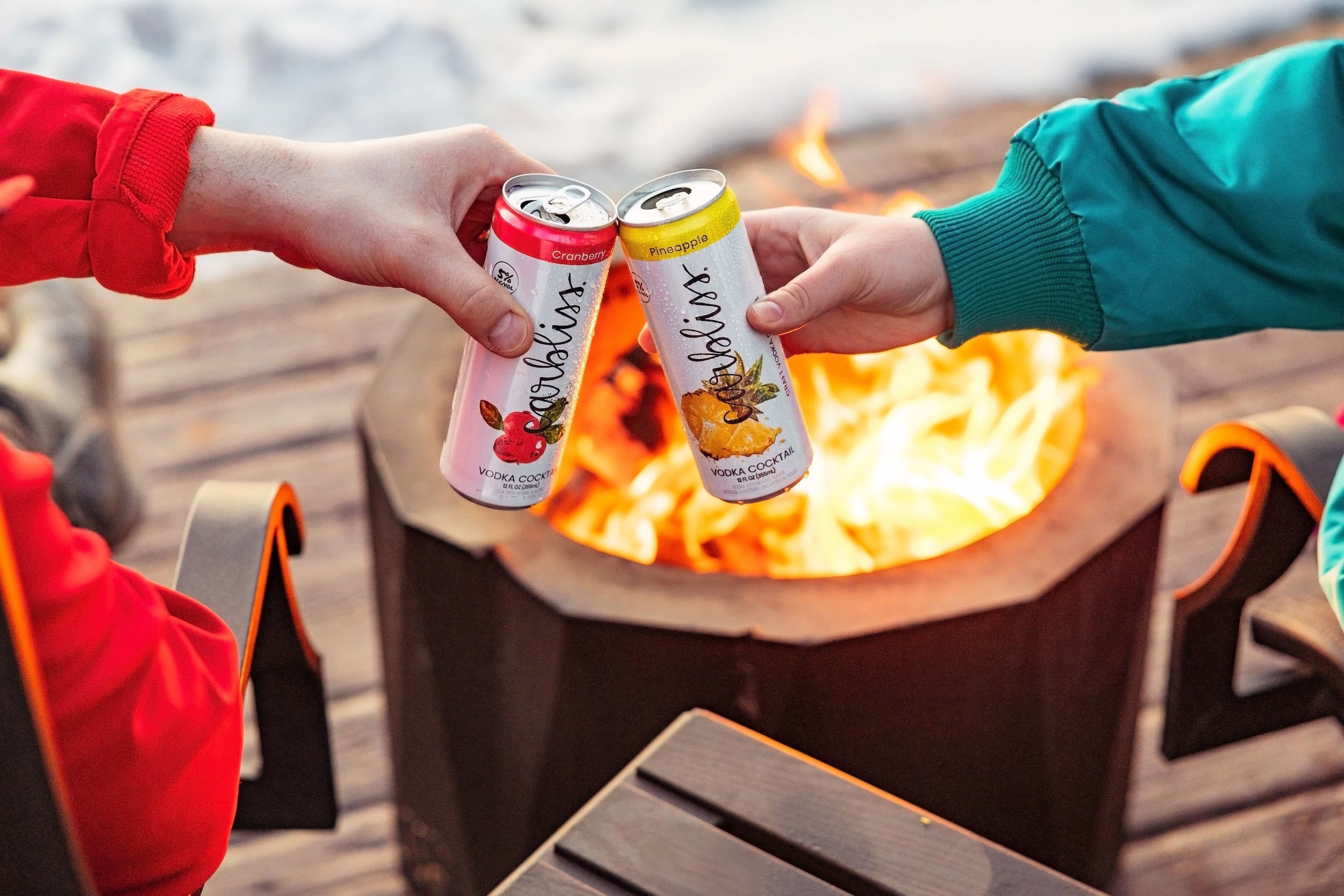 Two people holding cans of Anheuser-Busch InBev products near a fire pit.