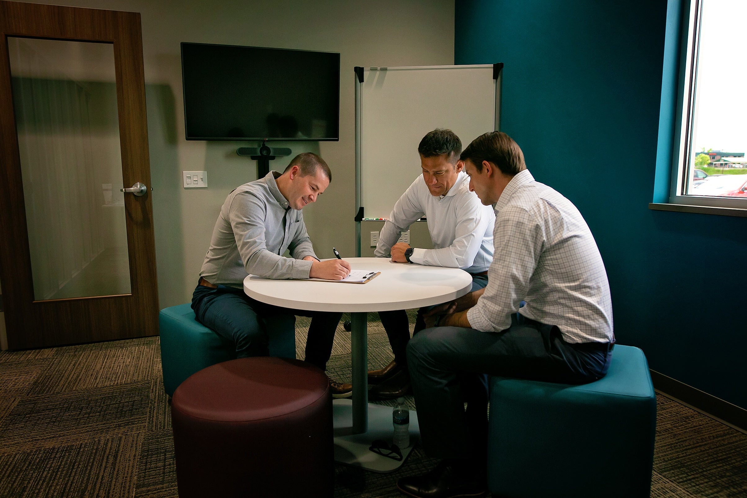 Three men in a modern office meeting room discuss documents around a white table.